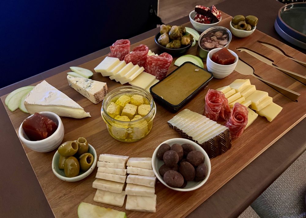 Charcuterie board consisting of: apple slices, caramelized onion cheddar, olives, quince paste, Brie, blue cheese, white cheddar, manchego, goat cheese, chocolate covered cherries, duck pate, soppressata, gruyere, octopus, emmental, dark chocolate raspberry pistachio bark. 