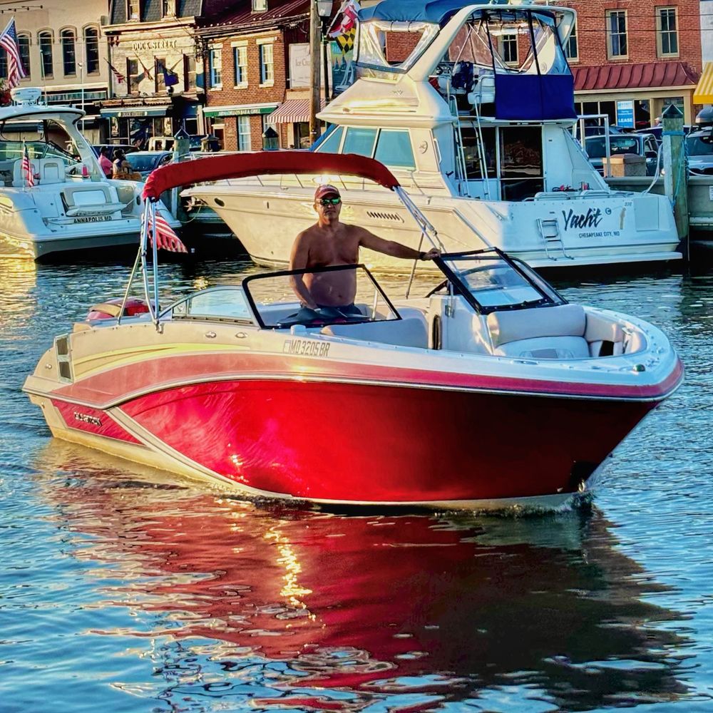 A shirtless man in a cap operates a striking red and white motorboat in calm harbor waters during golden hour. The boat’s glossy red hull creates vivid reflections on the water’s surface. In the background, brick buildings line the waterfront with other boats moored at the marina, creating a classic Annapolis Harbor scene.