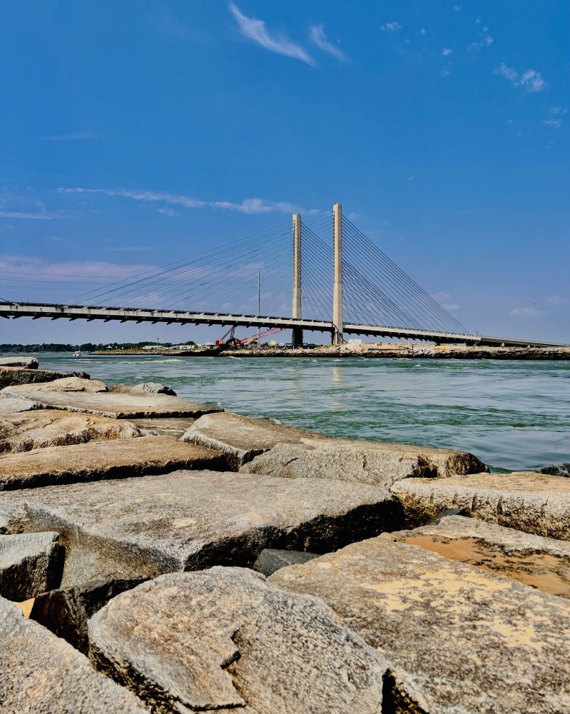 Weathered concrete breakwater rocks in the foreground frame a view of the Charles W. Cullen Bridge (Indian River Inlet Bridge,) a white cable-stayed bridge spanning turquoise waters under a bright blue sky with wispy clouds. The rocky barrier extends along the shoreline at Delaware Seashore State Park's Fisherman's Walkway. 
