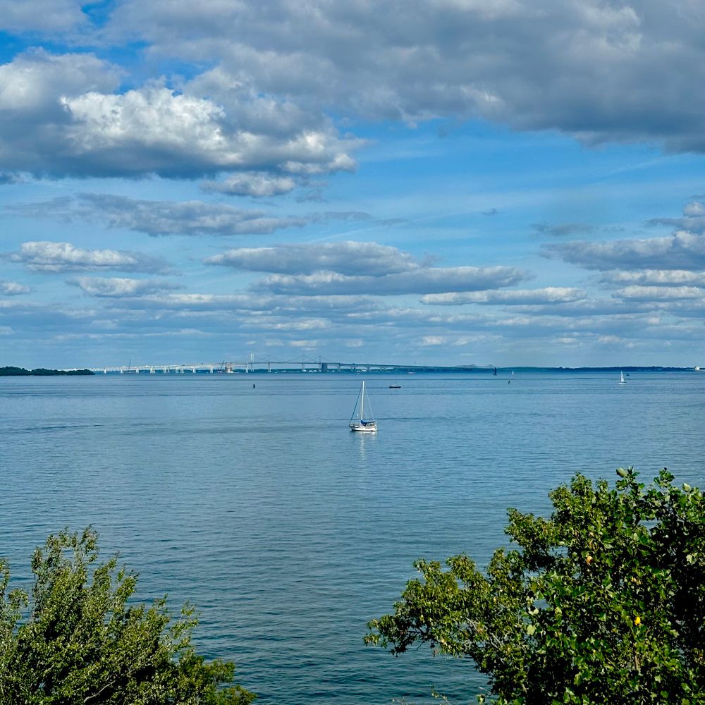 Sailboat on calm Chesapeake Bay waters with bridge spanning horizon under cloudy blue sky. Annapolis, Maryland. (U.S.) 
