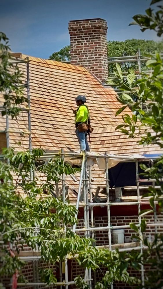 Construction worker in bright yellow safety shirt and hard hat stands on scaffolding while installing wooden shingles on the steep roof of a brick building with a prominent chimney.