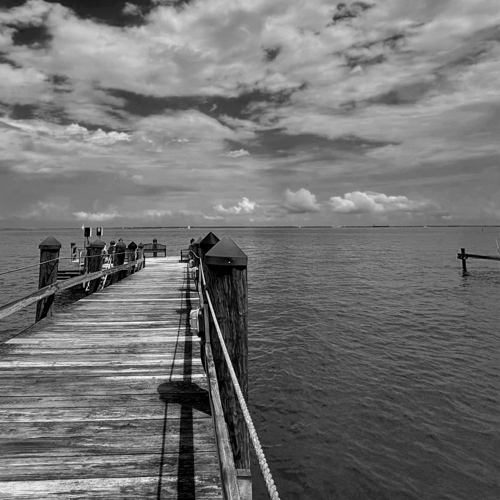 A black and white photograph of a weathered wooden pier extending into the calm waters of the Chesapeake Bay. The pier features rustic railings and small structures, leading the eye toward the distant horizon where sailboats dot the water. Dramatic clouds fill the expansive sky above the serene waterscape, creating a contemplative mood perfect for a morning walk and quiet reflection. Annapolis, Maryland. (U.S.) 📷 07/25/25.