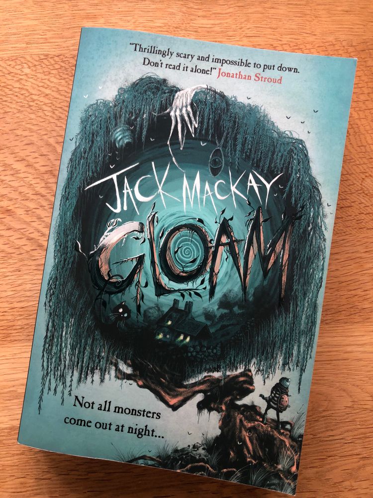 Front cover of ‘Gloam’ by Jack MacKay showing a girl with a backpack at the base of a willow tree. In the centre of the tree is a swirling vortex featuring a small house and a black cat. A skeletal hand with long, thin fingers is clawing the top of the tree, and an amulet with a picture of an eye is dangling from the tree’s branches. The tagline reads, ‘Not all monsters come out at night…’ (Illustrated by Ben Joel Price) 