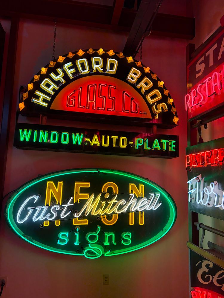 Two neon signs: HAYFORD BROS. (white in a, arc over) GLASS CO (red & inside the arc) WINDOW (green) AUTO (yellow) PLATE (green); Gust Mitchell (white, diagonal & in cursive) NEON (yellow with letters interrupted by the cursive neon, signs (in green at the bottom of the green outlined oval)