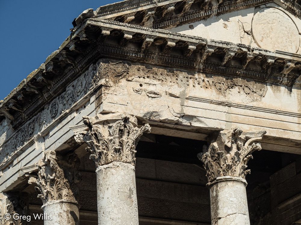 Detail of the Temple of Augustus in Pula. A well-eroded triangular pediment, cornice, & frieze are supported by Corinthian capitals atop plain columns. Blue sky in the upper left.
