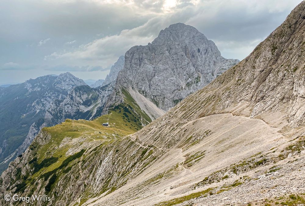 A gray limestone mountainside of Brana descends from the top right. Two trails through the rocky scree meet the mountain ridge right where it breaks -- a saddle green with grass and small shrubs. In the center of the saddle lies the hut. The peak of Planjava rises at back. Clouds fill the sky