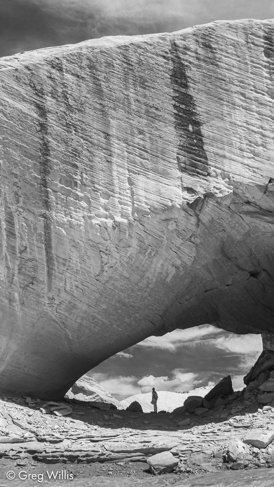 A black and white image. A lone figure stands in shadow underneath a very thick sandstone arch, with diagonal striations and dark vertical streaks of desert varnish. Above and below the arch, a dark sky with clouds 