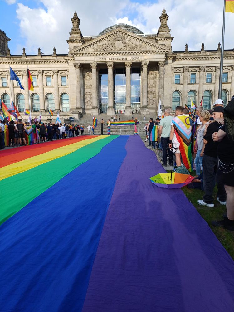 Riesige Regenbogenflagge, ausgebreitet vor der Freitreppe zum Reichstagsgebäude, gesäumt von vielen bunt gekleideten Menschen 