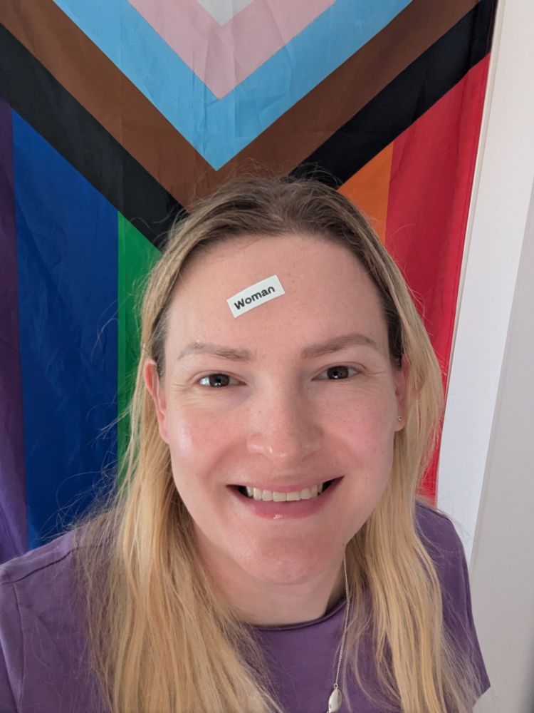 Chris Northwood, a blonde trans woman smiles into the camera stood in front of a progress pride flag. A label with the word woman is stuck to her forehead.