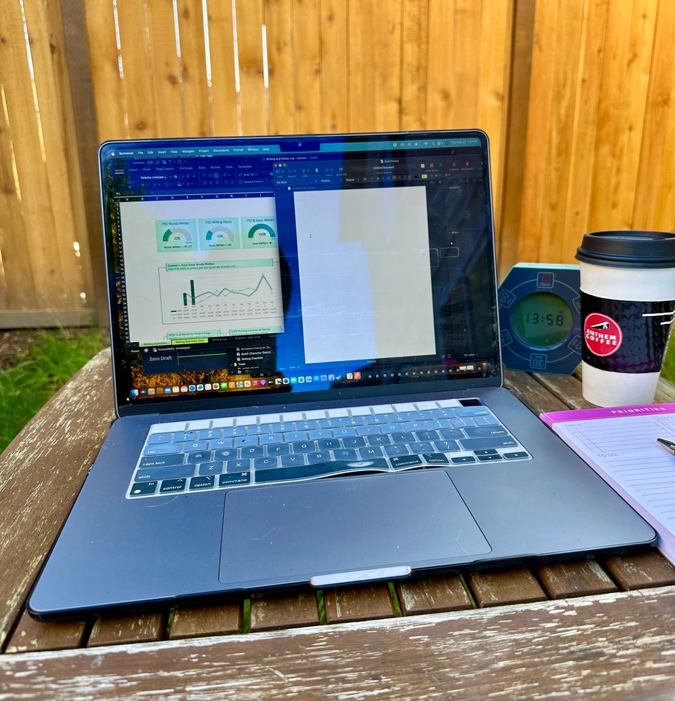 An open laptop sitting on a weather-worn wooden table in a weedy backyard and a cedar fence in the background. There's a blank Scrivener page on the laptop screen itching for some words. To the right of the laptop is a little timer clock, a to-go coffee cup, and a paper notepad.