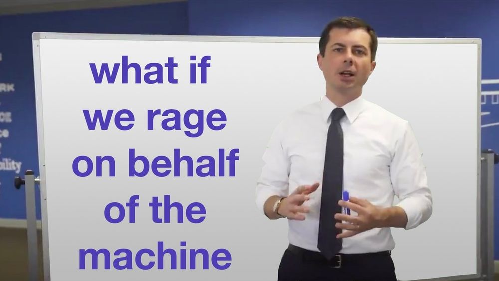 Pete Buttigieg, wearing a white shirt black pants and tie, stands in front of a white board speaking. Written on the whiteboard is the phrase "what if we rage on behalf of the machine"