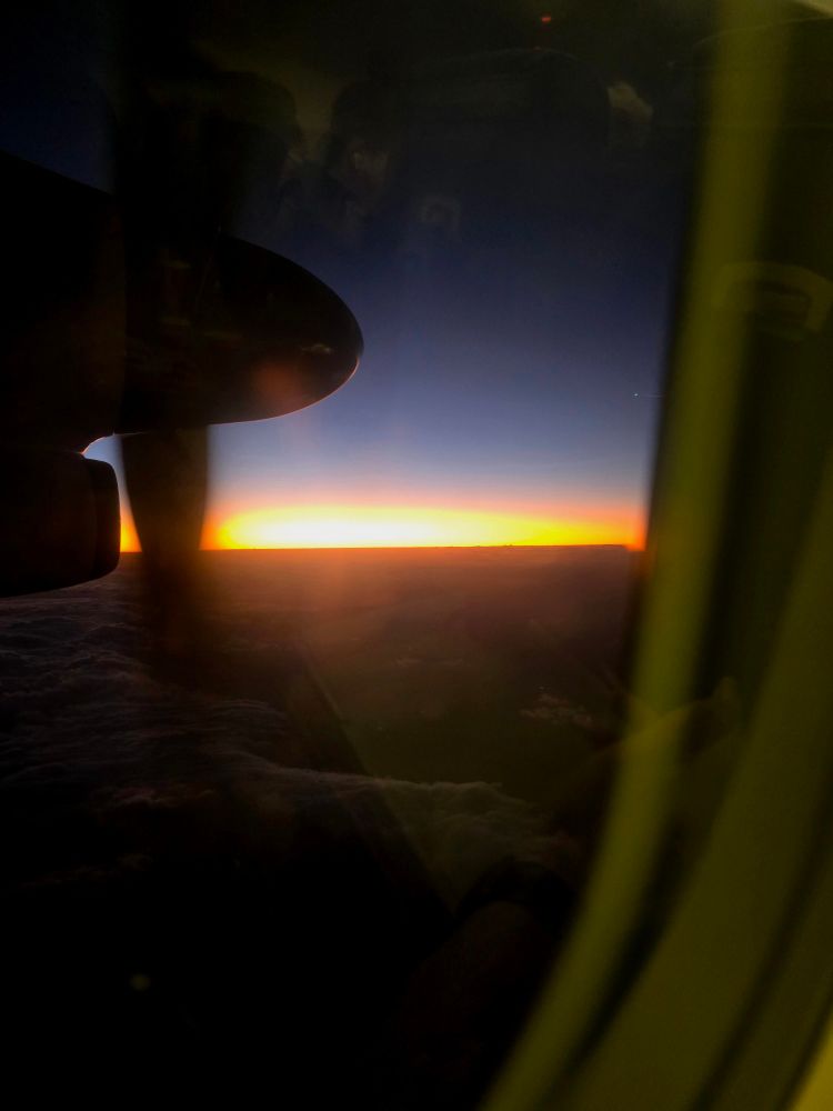 A window view from inside a DASH 8 airplane flying over cloud cover with the spinning prop just to the left of the setting sun. The sky's colour gradient from top to mid-picture going black to purple to blue and then a firey susnet and the dark grey-black cloud underneath.