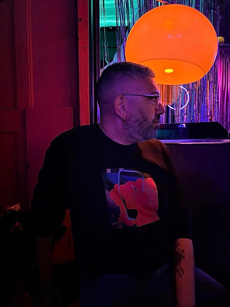 Photo de moi de profil, dans le bar Candy Darling à Barcelone. Je porte un sweat noir(avec un dessin que je suis toujours incapable d’expliquer quand on me dit « c’est beau c’est quoi ? ». Je suis éclairé par une lampe orange tout droit sortie des années 70
