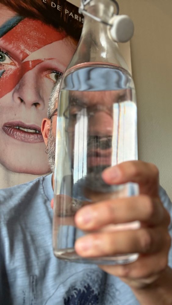 Selfie débile dans lequel je tiens une bouteille d’eau devant mon visage qui est donc déformé. Derrière moi, l’affiche de l’expo « David Bowie is » à la philharmonie. 