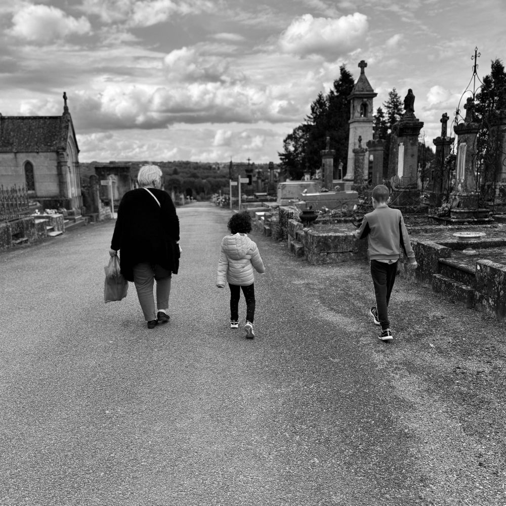 Photo en noir et blanc au format carré. Ma mère descend l’allée d’un cimetière, avec deux de ses petits enfants qui marchent à côté d’elle. Au milieu des tombes. Sous un ciel nuageux. Son visage semble tourné vers eux. 
