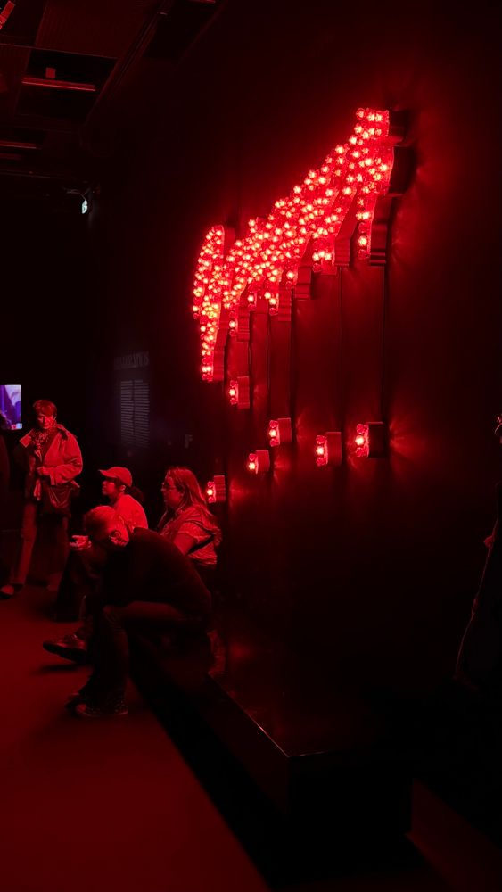 Photo de l’expo DISCO à la philharmonie, sombre avec le mot FOREVER écrit avec des ampoules rouge sur un mur à droite. En-dessous, des personnes assises sur un banc. La photo baigne dans une lumière rouge sombre 