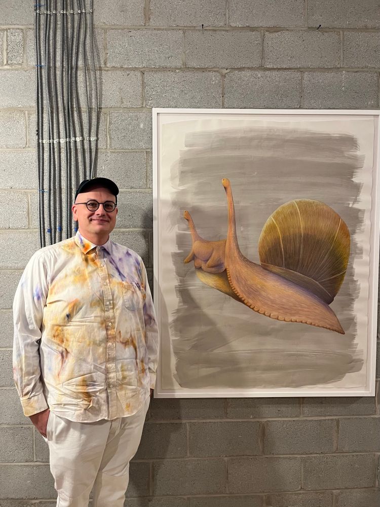 me, wearing an ice dyed white dress shirt, black cap, and white pants, next to my wax crayon and ink on paper drawing of a snail