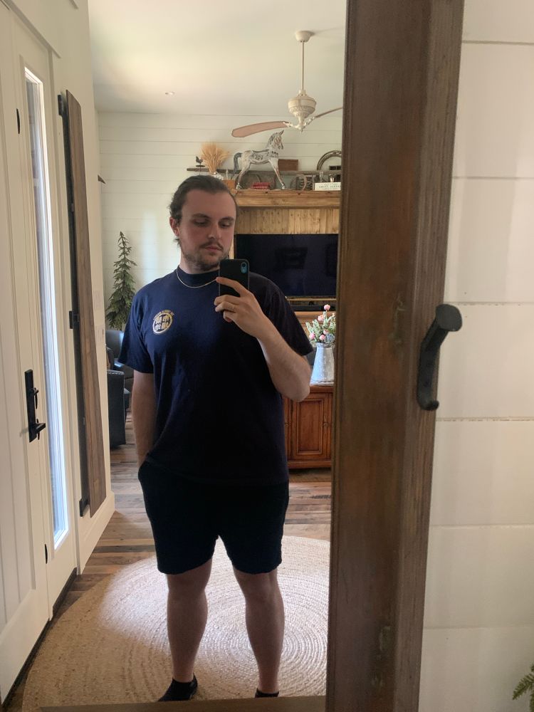 A full length mirror selfie of me in a blue shirt and black shorts. I’m wearing a small gold chain necklace and am looking at my phone.