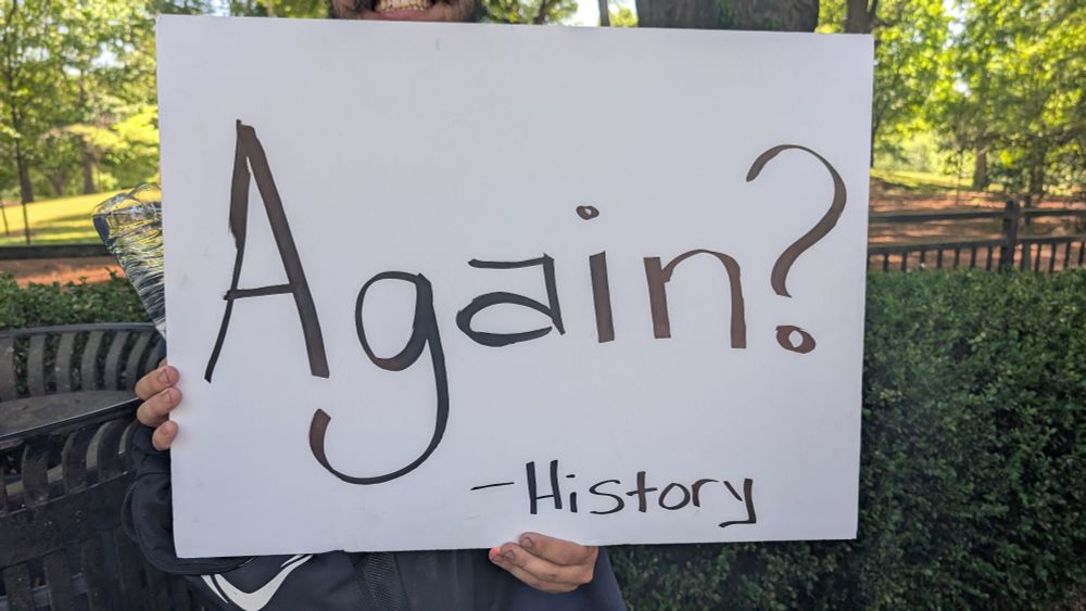 A protest sign that says "Again? -History". Someone is holding it up but you can't see their face. 