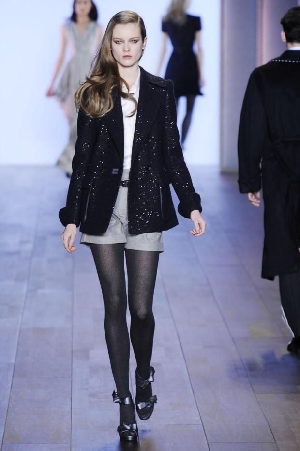 A female model walks down a runway at a fashion show in gray shorts, white shirt, dark blue tights, high heels with an ankle strap and wearing a midnight blue sparkly boucle peacoat.