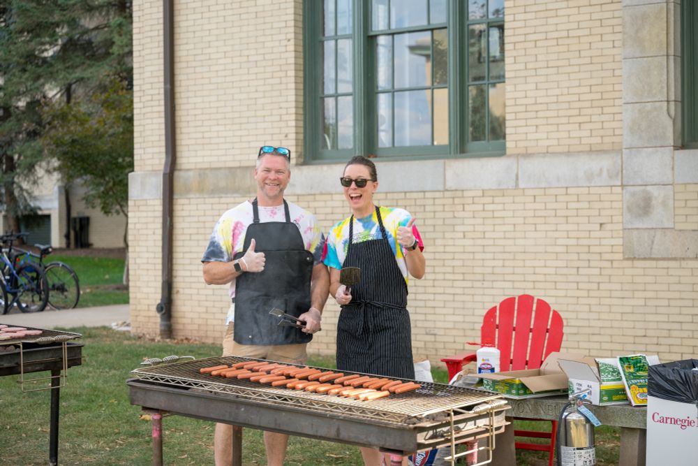 Ken Hovis and Maggie Braun smile at the camera as they stand next to a grill with hotdogs on it.