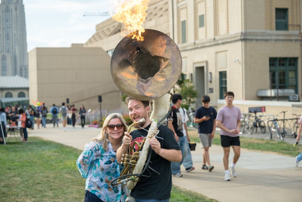 Dean Barbara Shinn-Cunningham and Eric Yttri smile at the camera. Yttrium holds a flaming sousaphone.