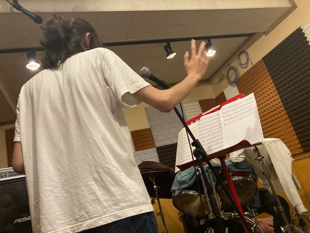 録音スタジオの風景その2。一枚目とは別のスタジオで、歌の録音をしている。一本のマイクの前に男性が立っている。マイクの後ろ側には譜面台が立ててあり、楽譜が置かれている。