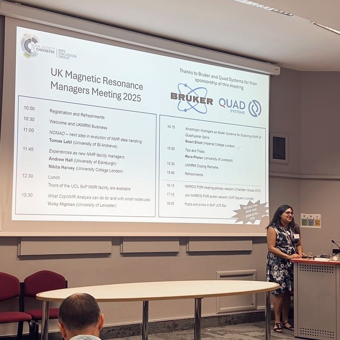Nikita standing in front of a projector screen showing the agenda for the UK Magnetic Resonance Managers Meeting 2025