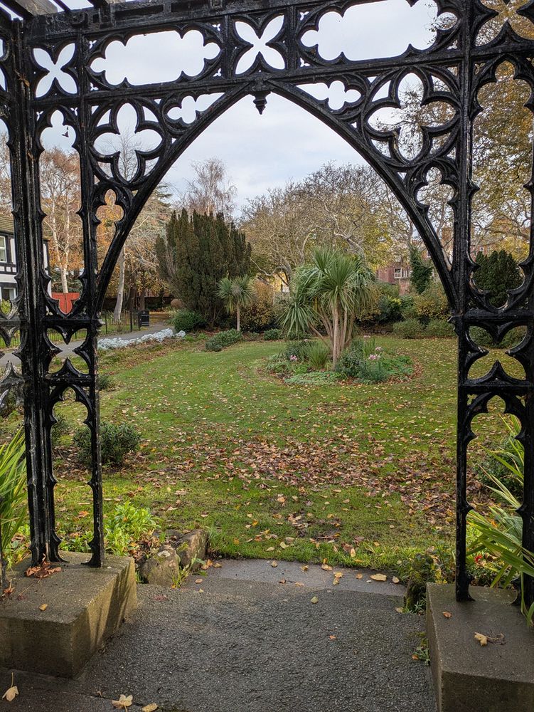 A decorative black wrought iron archway frames a leafy garden path with scattered autumn leaves and trees in the background.