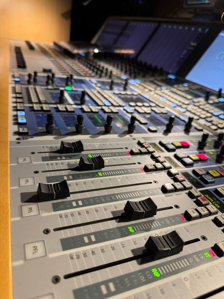 Blick auf ein Tonmischpult von schräg rechts. Zu sehen sind Fader in unterschiedlichen Stellungen, Taster und Drehregler. Zudem Kontrolldisplays.