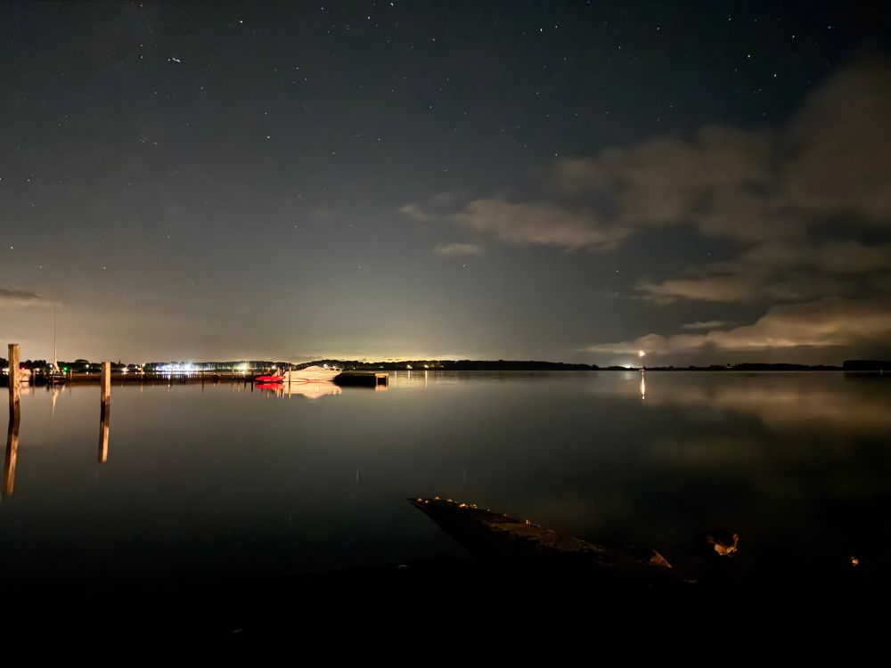 In der Nacht leuchtet fern ein Leuchtfeuer. Vorne am Steg ist ein Motorboot vertäut.
Das Wasser ist spiegelglatt, Sterne und Wolken stehen am Himmel.