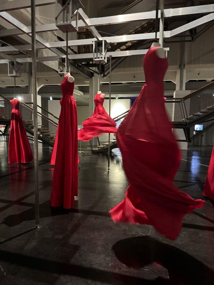 An mehreren Torsos hängen lange rote ärmellose Kleider. Die Torsos hängen an Stangen, die nacheinander durch Elektromotoren schnell gedreht werden. Eine Zeitlang laufen sie gemeinsam, um dann gemeinsam zur Ruhe zu kommen.