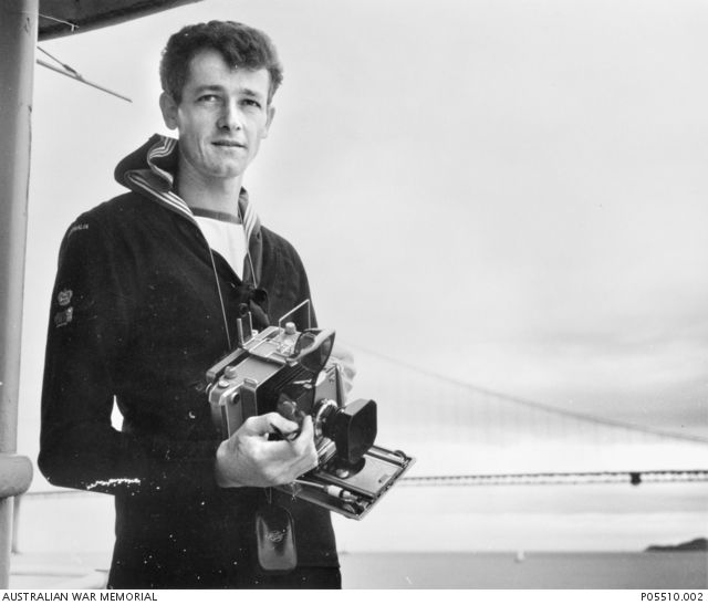 Informal portrait of Leading Airman Photographer (LAPHOT) Richard John (Rick) Reynolds, on board a tug in San Franciso harbour. He is using a Linhoff Technica camera to take photographs of his ship, HMAS Perth (II), as it sails under the Golden Gate bridge. LAPHOT Reynolds was a survivor of the sinking of HMAS Voyager on the night of 10 February 1964, the result of a collision with HMAS Melbourne during exercises off Jervis Bay, southern NSW.