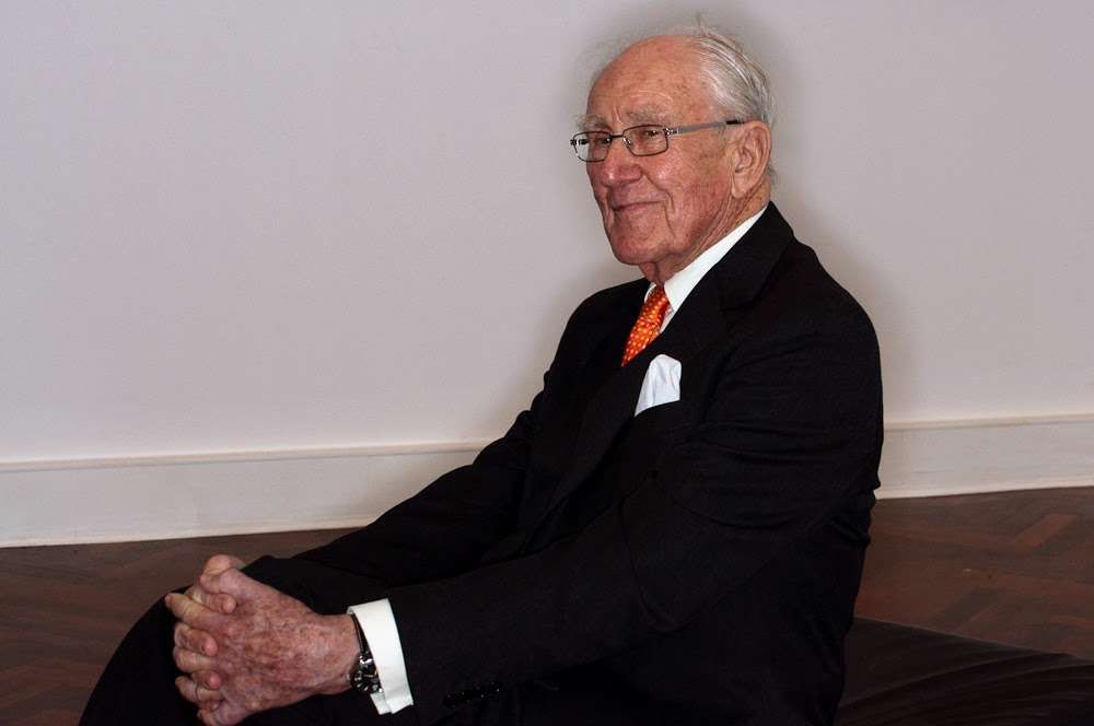 A very well dressed elder gentleman is seated facing to your left and clasping his hands around one knee.