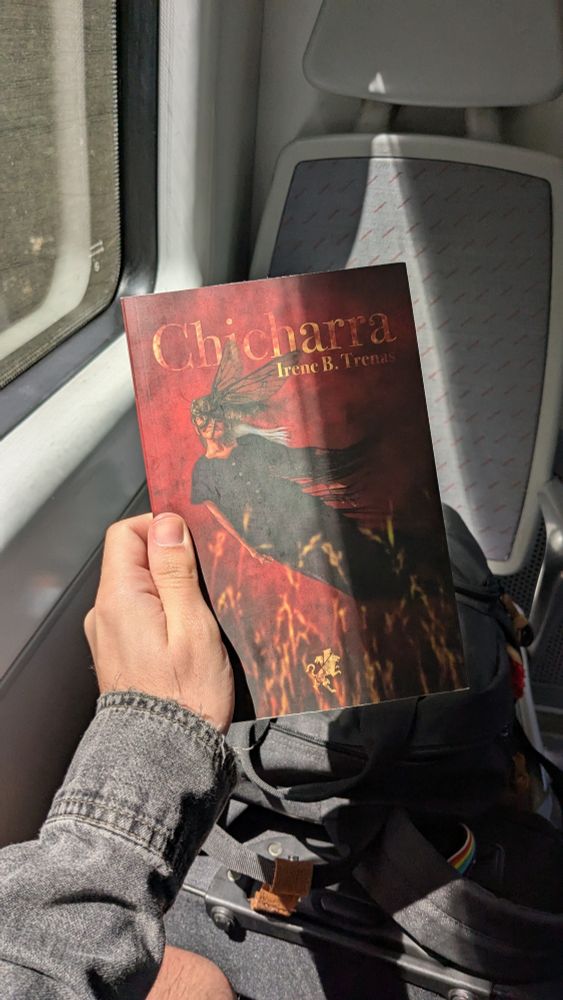 Foto del libro frente al asiento gris de un tren de cercanías. Se ve un trocito de ventana. La mano que sujeta el libro lleva una camisa vaquera hasta la muñeca. Sí, la mano, por qué no. El libro está iluminado a medias por el sol que entra a través del cristal. Es rojo, en él, una chicharra gigante se agarra a la cabeza una anciana, lo vemos desde nuestra diminuta posición entre haces de hierba amarilla y seca. 