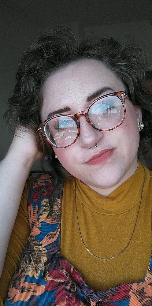 white female with salt and pepper hair wearing a mustard turtleneck and patterned vest and glasses smiling