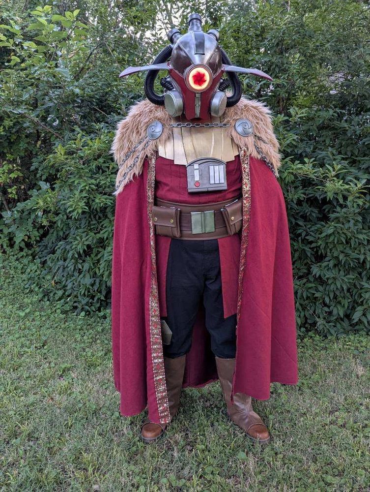 A full body shot of me in my mostly-finished Marchion Ro costume. I am standing straight up in front of dense bushes, wearing black cargo pants with hexagonal silver knee pads, mid-calf brown boots, a layered brown belt with a chrome buckle and two large square pouches, and a red shirt with vertical pin tucks around my entire torso and a wasit cape of the same red linen extending from the belt to just above the knees. Over all this is a segmented golden armor plate covering my upper chest and long red cape covering my arms with a tan faux fur mantle over the shoulders and silver chains hanging around the back. On my head is an elaborate red and silver helmet with black rubber hoses looping around to imply an air filtration system, wide sweeping fins protruding from the left and right sides, and a glowing red lens in the middle of the face with the dark outline of a hurricane at its center