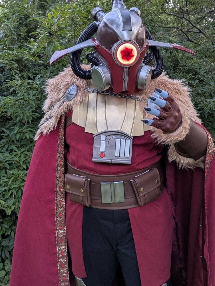 A knee-up shot of me in my mostly-finished Marchion Ro costume. I am standing straight up in front of dense bushes, wearing black cargo pants with hexagonal silver knee pads, mid-calf brown boots, a layered brown belt with a chrome buckle and two large square pouches, and a red shirt with vertical pin tucks around my entire torso and a wasit cape of the same red linen extending from the belt to just above the knees. Over all this is a segmented golden armor plate covering my upper chest and long red cape covering my arms with a tan faux fur mantle over the shoulders and silver chains hanging around the back. On my head is an elaborate red and silver helmet with black rubber hoses looping around to imply an air filtration system, wide sweeping fins protruding from the left and right sides, and a glowing red lens in the middle of the face with the dark outline of a hurricane at its center. My left hand is raised in a grasping motion, revealing blue-grey finger tips with long sharp black nails, a fingerless leather glove, and a leather vambrace trimmed in the same fur as the cape.