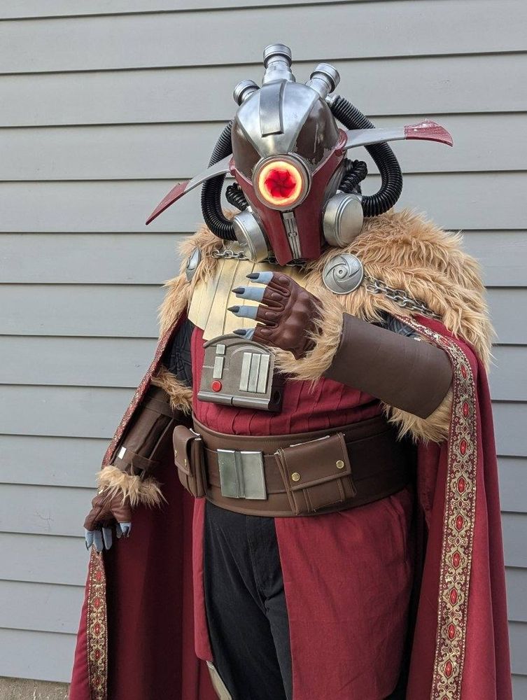 A knee-up shot of me in my mostly-finished Marchion Ro costume. I am standing straight up in front of a grey wooden wall, wearing black cargo pants with hexagonal silver knee pads, mid-calf brown boots, a layered brown belt with a chrome buckle and two large square pouches, and a red shirt with vertical pin tucks around my entire torso and a wasit cape of the same red linen extending from the belt to just above the knees. Over all this is a segmented golden armor plate covering my upper chest and long red cape covering my arms with a tan faux fur mantle over the shoulders and silver chains hanging around the back. On my head is an elaborate red and silver helmet with black rubber hoses looping around to imply an air filtration system, wide sweeping fins protruding from the left and right sides, and a glowing red lens in the middle of the face with the dark outline of a hurricane at its center. My left hand is raised in a grasping motion, revealing blue-grey finger tips with long sharp black nails, a fingerless leather glove, and a leather vambrace trimmed in the same fur as the cape. My right hand is held out to my side, pushing back the edge of my cape and revealing the matching glove and bracer. Small glimpses of my upper arms under the cape show blue-grey sleeves with light grey tattoo patterns 