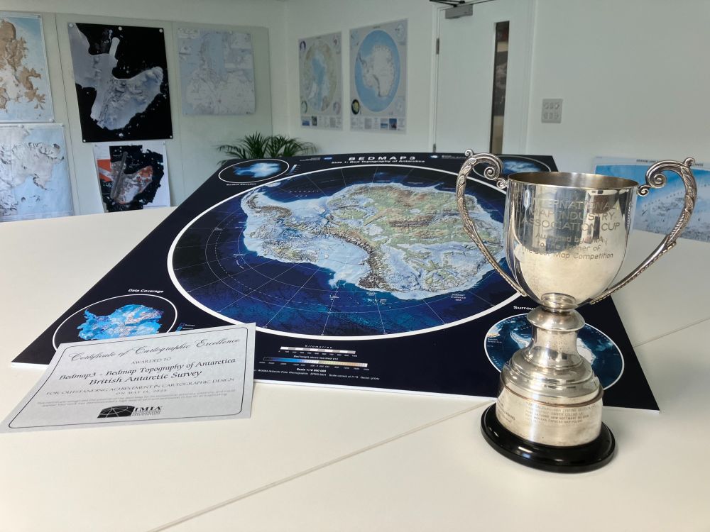 A photo of the trophy and certificate, with side A of the Bedmap 3 map shown behind. The map shows the bed topography of Antarctica underneath the ice.