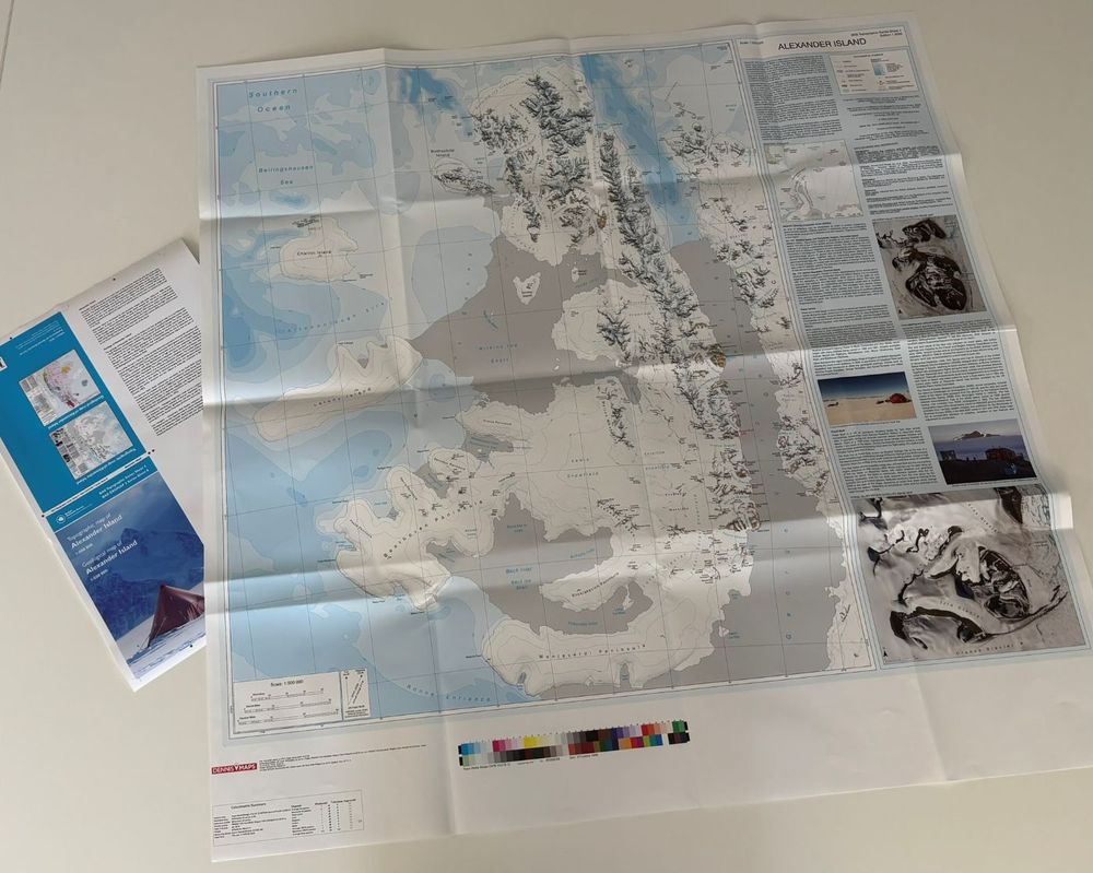 A photo of a map on a table. The map covers Alexander Island, a long island with large peninsulas coming off to the west. The land is white with contours and place names visible. Ice shelves are grey. Bathymetry is shown with intervals of cyan. A large information is shown to the right of the map with photos and text.
