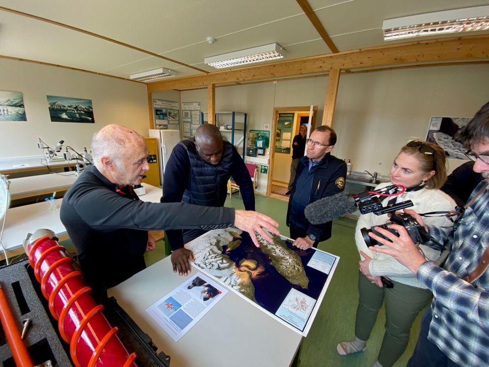 A photo of Dr Robert Mulvaney showing a map of of Ny-Ålesund and the Kongsfjorden region in Svalbard to visitors at the NERC Arctic Office. The visitors include the UK Foreign Secretary Rt Hon David Lammy and his Norwegian counterpart Espen Barth Eide. The photo shows 5 people stood around a map laid on a table, with one man filming.