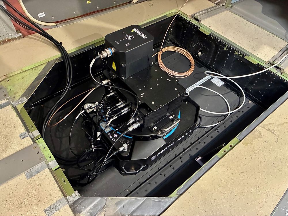 A photo of the aerial camera set up in the floor of the plane. The words 'applanix' and 'SOMAG' can be seen. There are lots of cables coming out of black boxes.