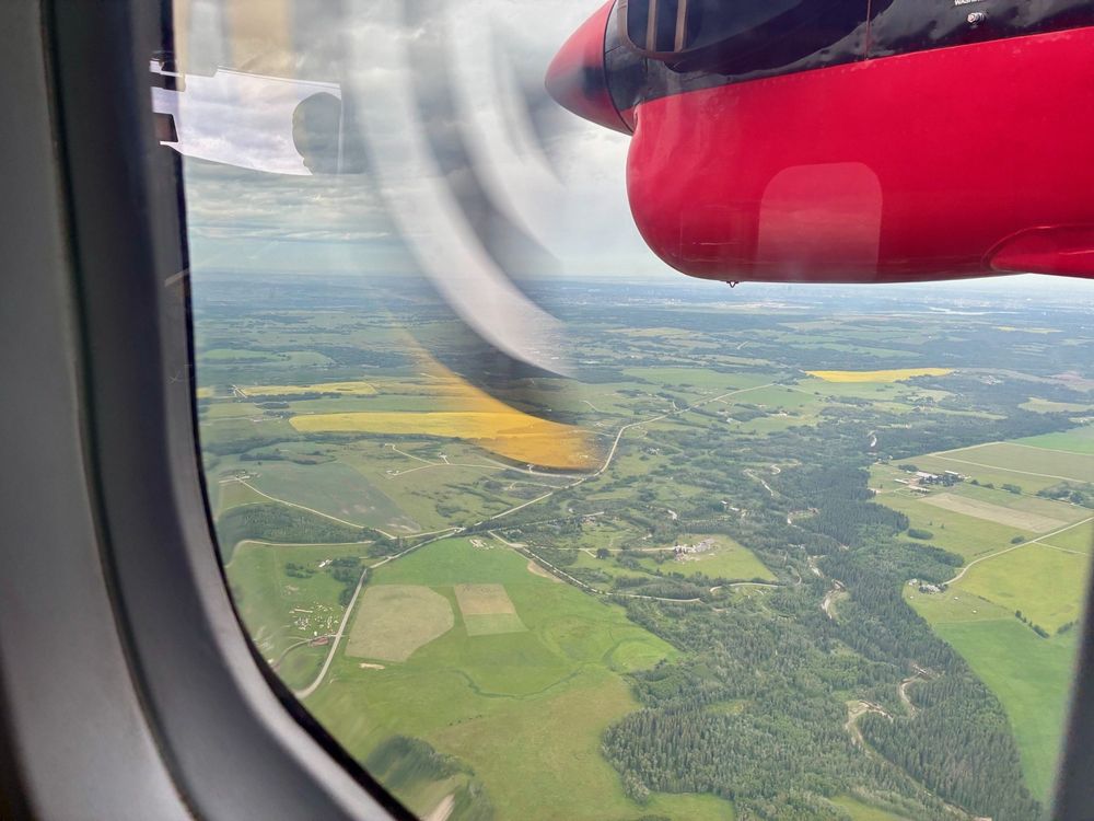 A photo out of the window during a test flight in the Twin Otter. A black and white propeller blade can be seen in motion. The view on the ground shows green fields and forested areas. 