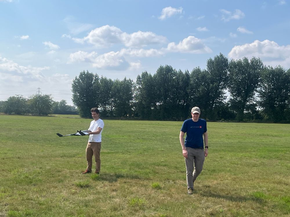 A photo of 2 men, one of them holding an ebee UAV, in a grassy field. There are trees in the distance and white fluffy clouds in a blue sky.