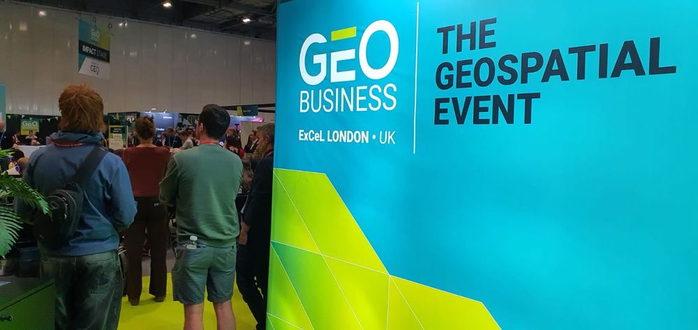 An image of a large banner saying "GEO Business ExCel London, UK. The Geospatial Event. There are people next to the sign listening to a talk.