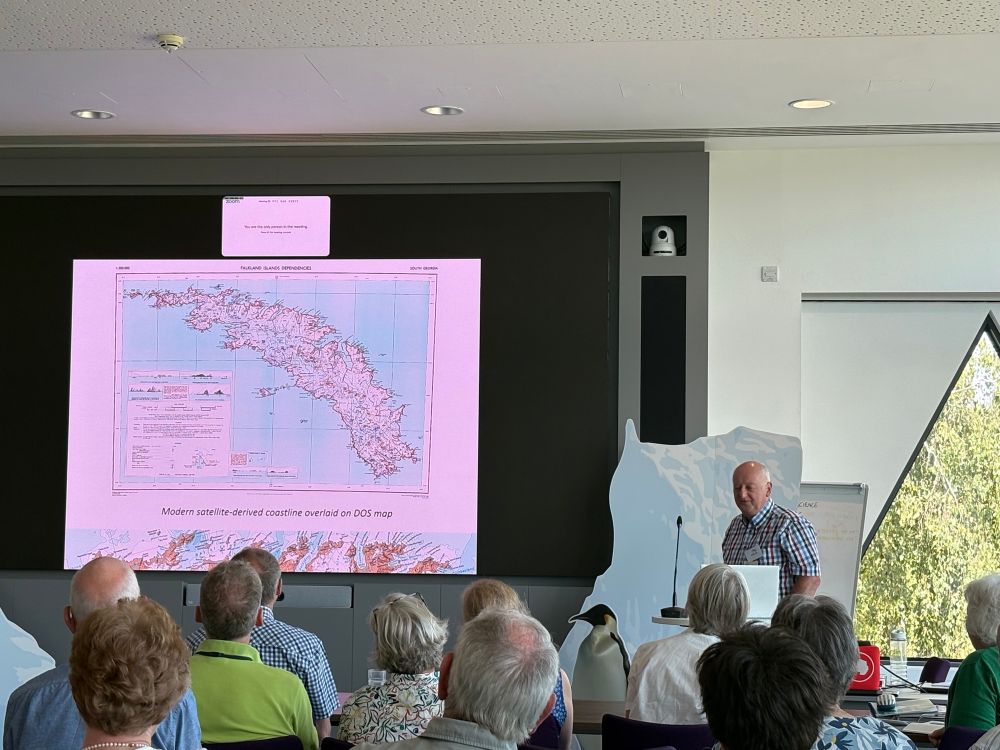 An image of a slide with a DOS map of South Georgia, with the modern coastline overlaid on the map. There are large differences in some places. The image shows many people watching the talk. 