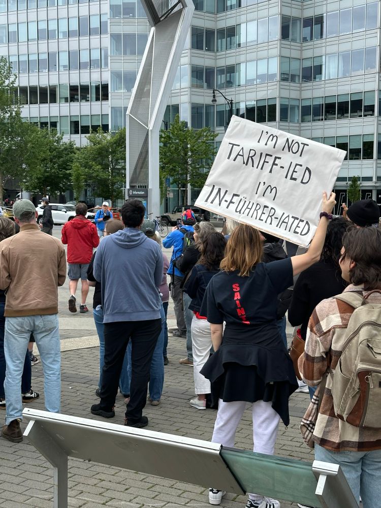 Photo of No Kings/No Tyrants protest in Vancouver, Canada. Shows people in a crowd facing the speaker and a person holding a protest sign that says “I’m not tariff-ied, I’m in-führer-iated”.