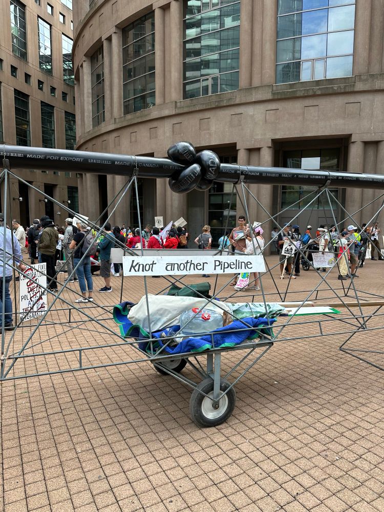 A protest in Vancouver, British Columbia called, Draw The Line, where in the photo someone has built a large structure on wheels that is a long, black piece of plumbing pipe tied into a knot in the middle. Below the pipe is a sign that says “knot another pipeline”.