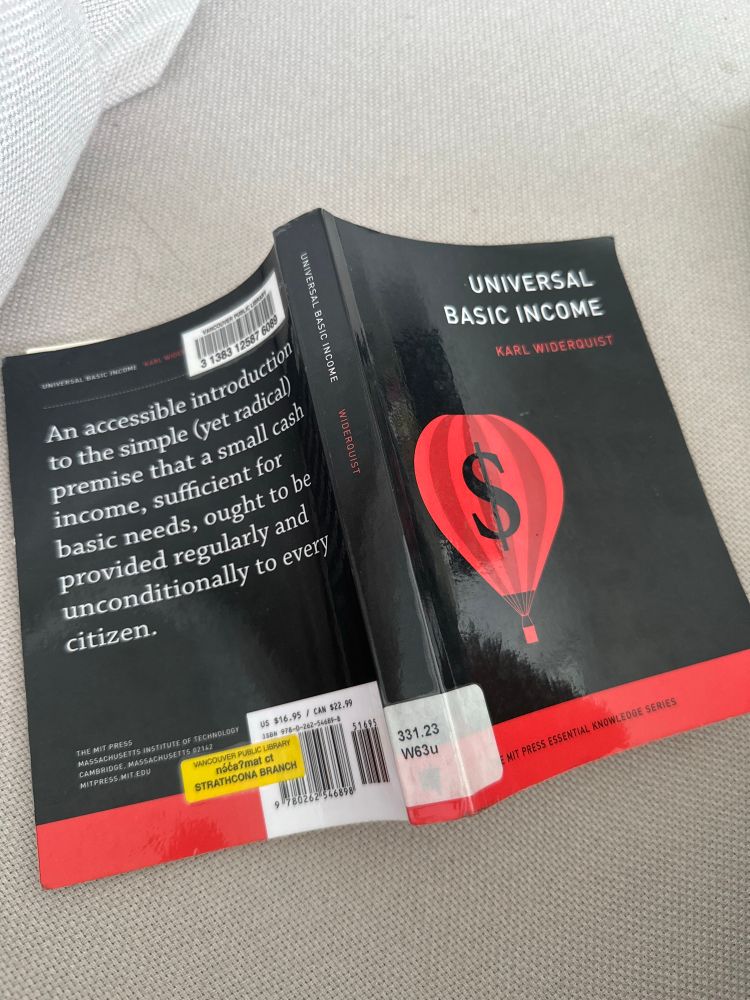 A paperback copy of Universal Basic Income by Karl Widerquist rests on a light-coloured fabric surface. The cover is black with a red hot air balloon featuring a dollar sign. The spine is slightly curved, indicating the book is in use. A yellow library sticker reads “Vancouver Public Library, on-demand at STRATHCONA BRANCH.” The back cover includes a quote describing the book as “an accessible introduction to the simple (yet radical) premise that a small cash income, sufficient for basic needs, ought to be provided regularly and unconditionally to every citizen.”